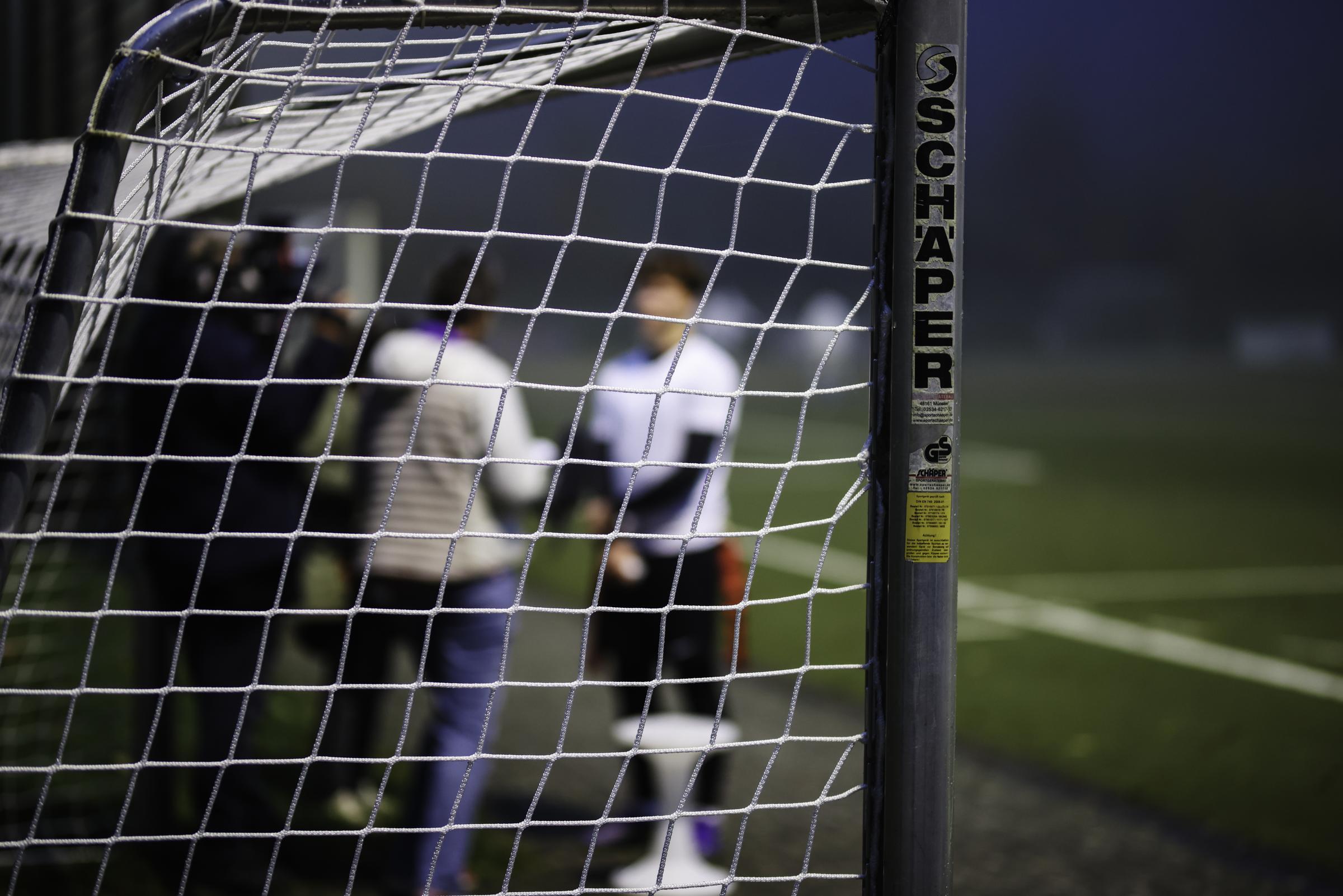 Auf dem Bild ist ein Sportplatz zu sehen, auf dem ein Fußballtor steht. Unscharf dahinter sind Jugendliche, die sich unterhalten. Foto: Martin Bondzio/Kindernothilfe Auf dem Bild ist ein Sportplatz zu sehen, auf dem ein Fußballtor steht. Unscharf dahinter sind Jugendliche, die sich unterhalten. Foto: Martin Bondzio/Kindernothilfe