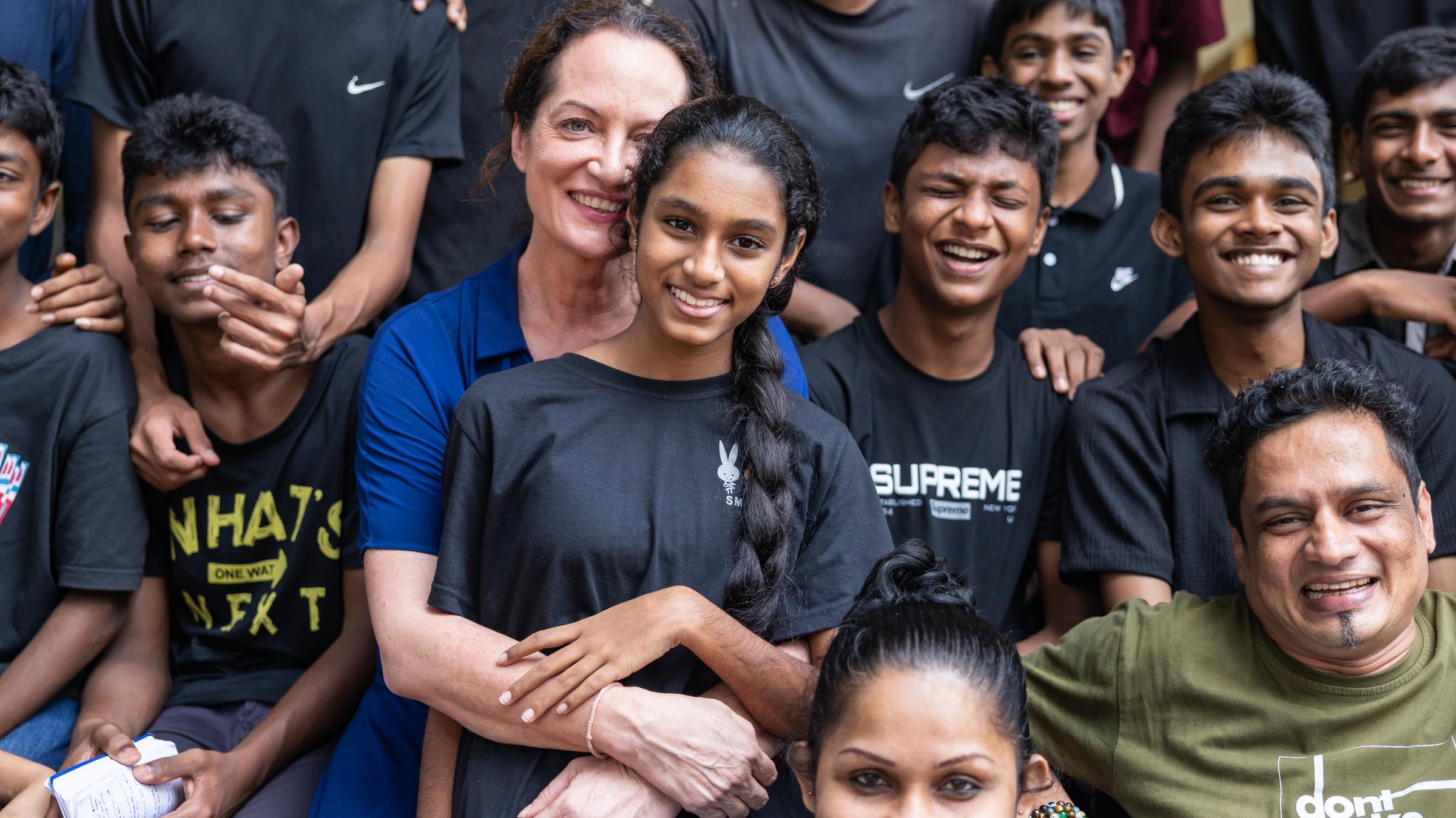 Schauspielerin und Kindernothilfe-Botschafterin Natalia Wörner umarmt ein Mädchen. Um sie herum sitzen weitere Jugendliche und Erwachsene. Alle schauen in die Kamera und lächeln. Foto: Sharni Jayawardena Schauspielerin und Kindernothilfe-Botschafterin Natalia Wörner umarmt ein Mädchen. Um sie herum sitzen weitere Jugendliche und Erwachsene. Alle schauen in die Kamera und lächeln. Foto: Sharni Jayawardena